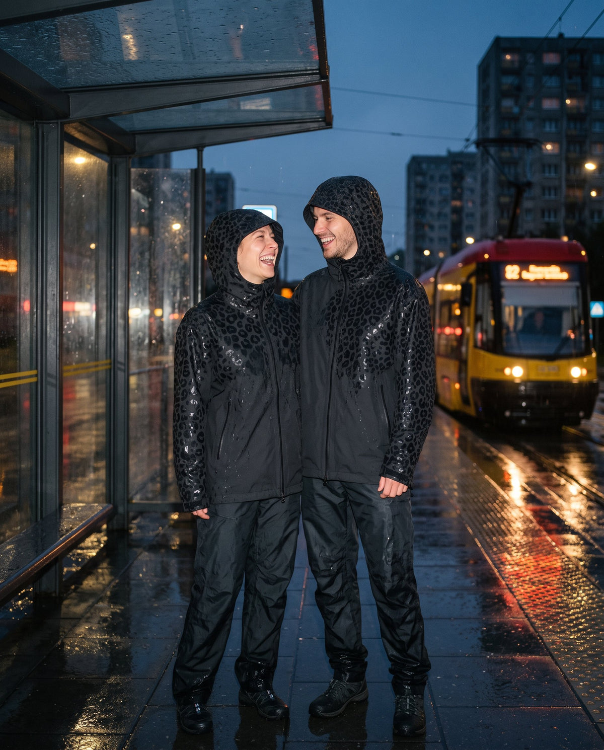 Para w kurtkach przeciwdeszczowych NIMBA śmieje się na przystanku tramwajowym w deszczu